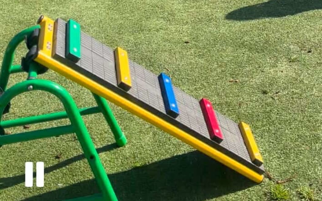 The play set is pictured on some grass in a fenced off area next to a carpark.