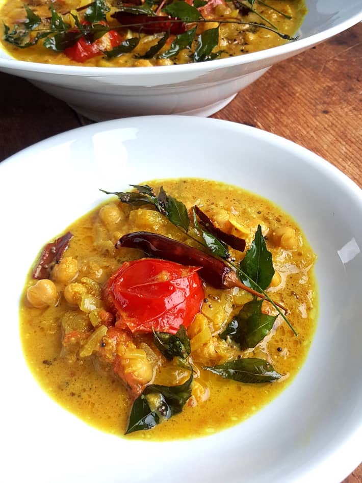A close-up image of coconut chickpea curry on a shallow white bowl, decorated with curry leaves and a tomato.