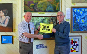 Richard and Graham shake hands. They are standing in the Mangaweka Gallery with paintings on the wall behind them. Richard presents Graham with the Gold Award.