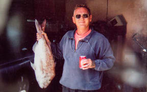 Archival photograph of a man (Ross Appelgren) holding a fresh caught fish in one hand and a can of beer in the other. He wears sunglasses and a blue sweatshirt and black shorts and smiles softly while looking at the camera.