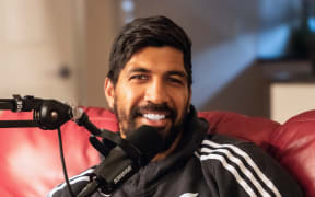 A bearded man wearing an All Blacks top smiles, in front of him is a microphone.