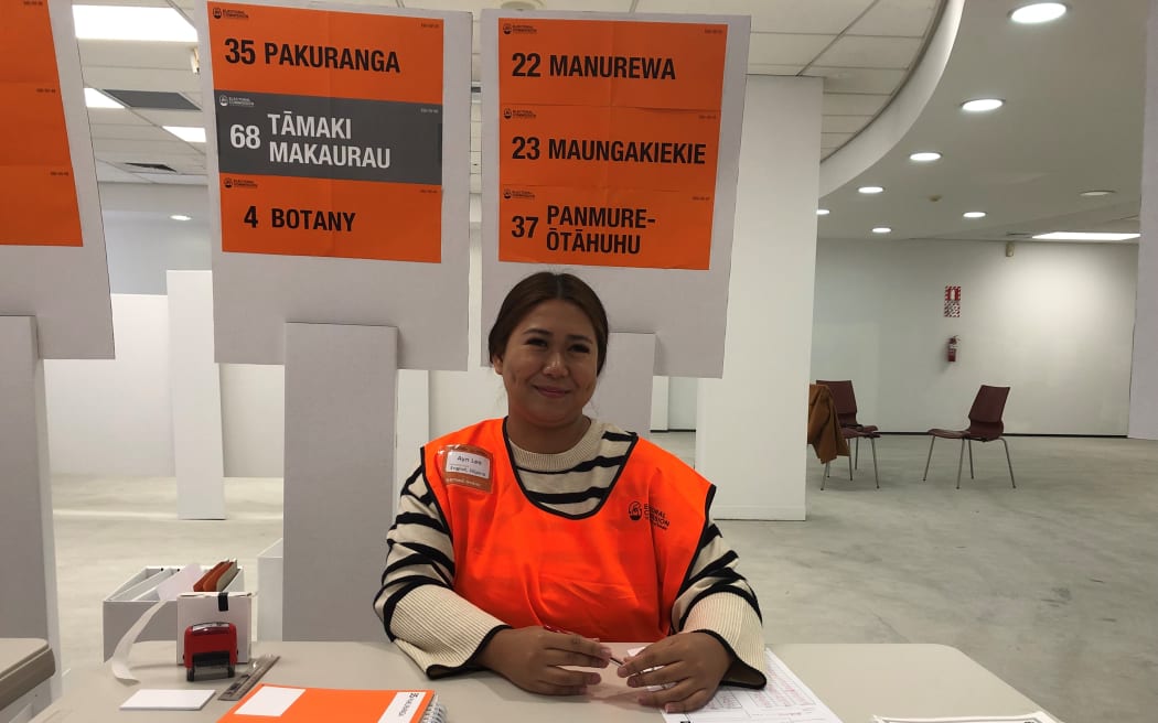 Ayn Lee smiles at the camera. She is sitting at a beige desk with electoral rolls placed by her side, wearing a fluorescent orange vest with her name on it. Behind here are official signs for guiding people to vote in the right queue: "35 - PAKURANGA", "68 - TĀMAKI MAKAURAU", "4 - BOTANY", "22 - MANUREWA", "23 - MAUNGAKIEKIE", "37 - PANMURE-ŌTĀHUHU".