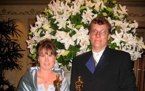 Tania and Richard stand side by side holding Oscars. They are both in fancy dress, Tania wearing a silvery blue ball gown and Richard wearing a black suit with satiny lapels.