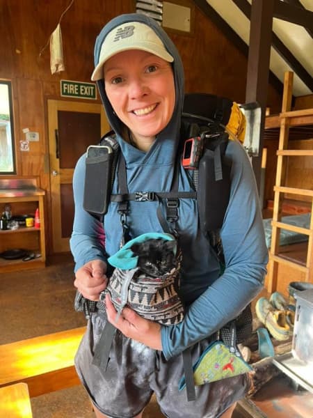 UK hiker Katie Evans in her gear at a hut with a black kitten in her shoulder bag.