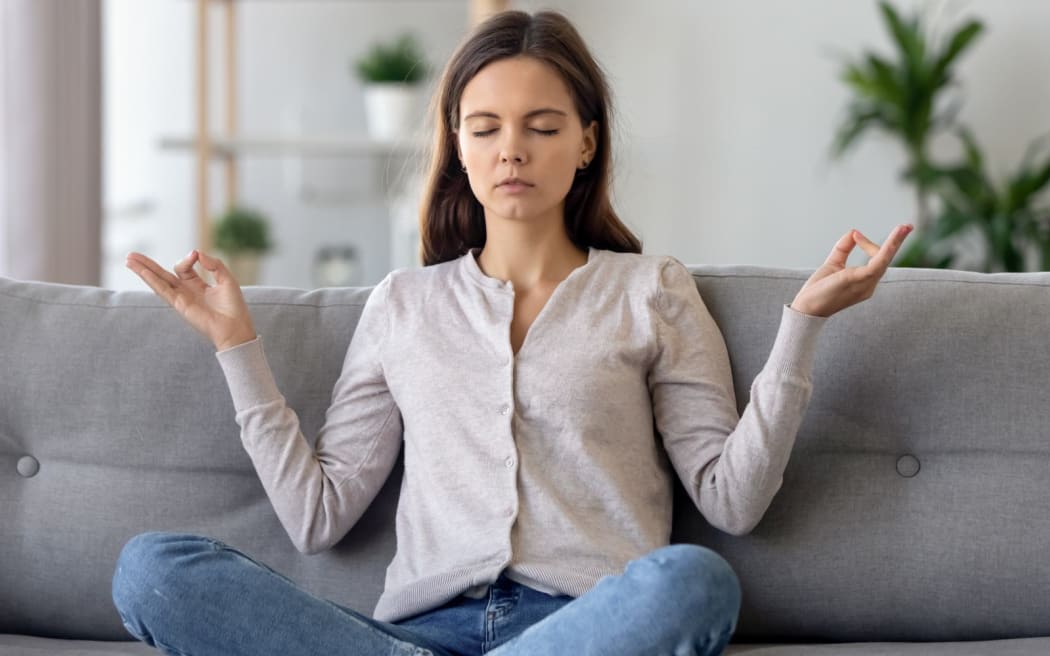 Peaceful young woman sitting in lotus pose on sofa in living room, meditating, attractive girl with closed eyes practicing yoga at home, stress relief concept, healthy good habit