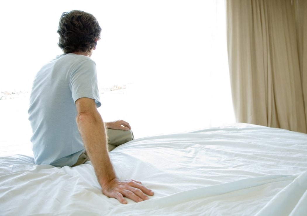 Mature man sitting alone on bed, looking out window (Photo by Eric Audras / AltoPress / PhotoAlto via AFP)