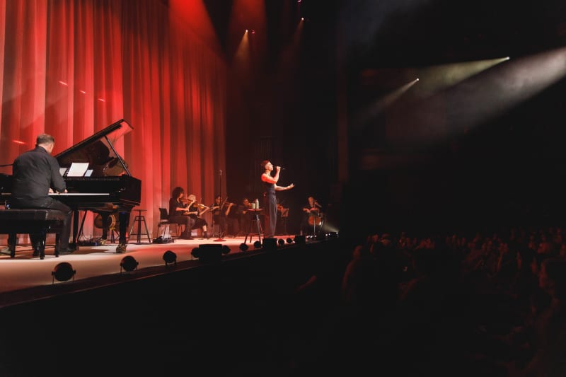 TEEKS, a muscular man in a black singlet and black jeans, sings into a microhpne with members of an orchestra and a red velvet curtain behind him.