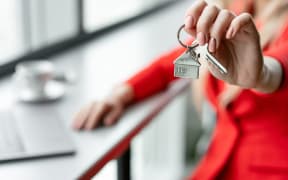 Mortgage concept. Woman in red business suit holding key with house shaped keychain. Modern light lobby interior. Real estate, moving home or renting property