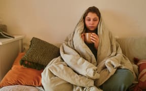Woman looking tired and sick, wrapped up in blanket on couch, holding a mug.