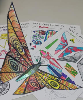 A yellow Awhi Creature hovering over a sample of designs for the creatures created by Papakura schoolchildren.