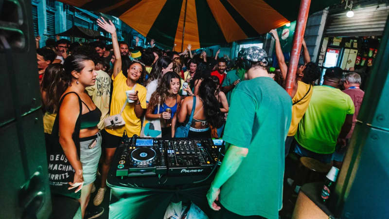 A group of colourfully dressed people surround a DJ on his decks.