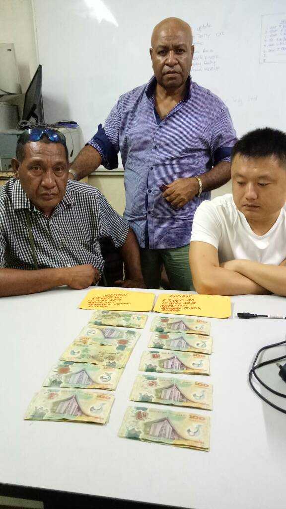 PNG Police Investigations Team Leader Senior Sergeant Apollos Terry (standing) with suspect Zhiyaun (Aaron) Cui (seated right) with the K10,000 cash allegedly offered to Senior Sergeant Terry (left)