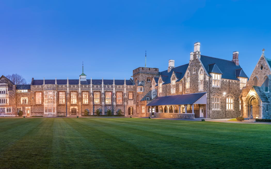 The campus of Christ's College. A large gothic building is lit from within, looking over a finely mown lawn. The evening sky is deep blue.