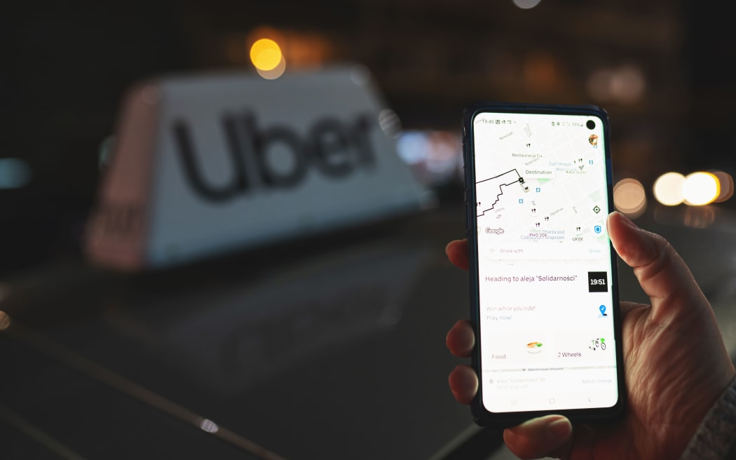 Uber app displayed on smartphone held in hand in front of Uber taxi sign on top of a car at night with city lights background with soft focus. Taking a cab concept. Warsaw, Poland - October 23, 2021