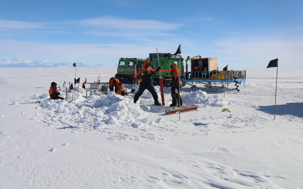 Four people in orange cold-weather gear engaged in various activities on a flat expanse of ice in front of a green-and-range Hagglund vehicle. Two people are wielding are long metal drill/pole, and the other two are crouched behind heaped up snow next to an array of flags. The sky is blue with a few bands of cloud.