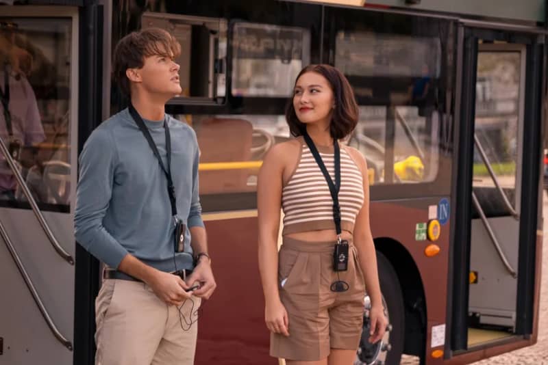 A young woman with a camera around her neck looks fondly at a young man with a bus behind them.