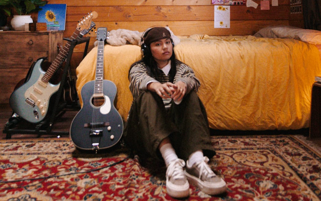 Paige is sat in what looks like a bedroom. It has cabin vibes. She is sat on a Persian Rug, the wall is wood paneled, and there are a couple of guitars leaning up against the bed,