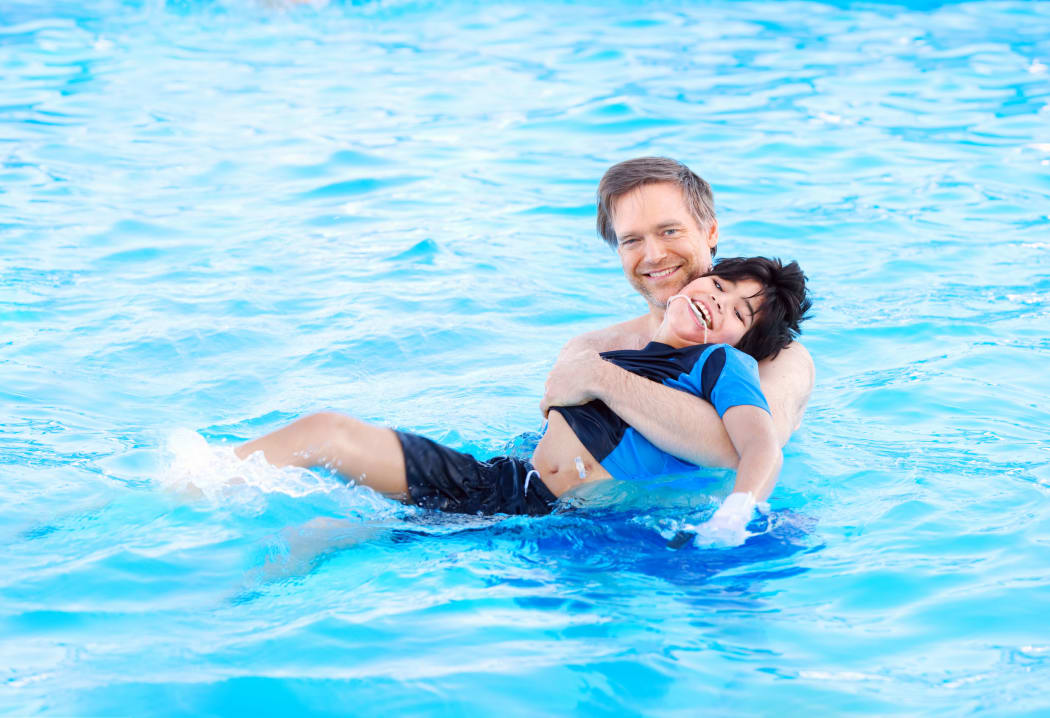 A photo of a father swimming in a pool with his biracial disabled son in his arms. The child has cerebral palsy.