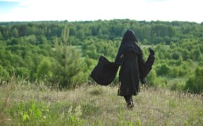 woman witch in a black cloak looks into the woods from a mountain covered with grass