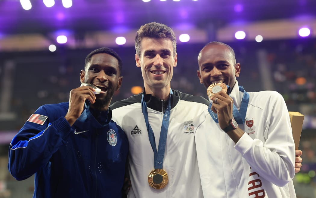 Three men stand arm in arm each with their medals around their necks. The silver and bronze winners have them in their mouths.