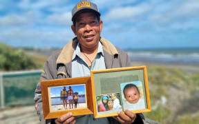 Faisal Syafii is smiling and wearing a cap while holding framed photographs of his family.