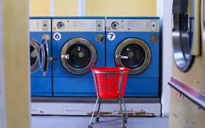 A red laundry trolley stands in front of a row of blue washing machines in a laundromat.
