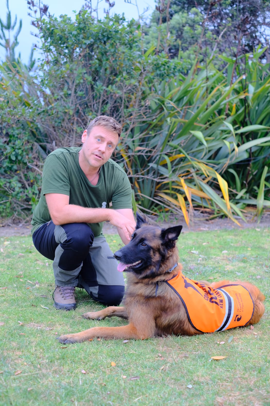 Fredrik kneels on the grass with Hawke, the dog, next to him, wearing an orange jacket.