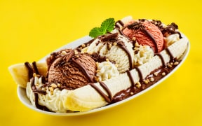 Tropical banana split with chocolate drizzle over three scoops of chocolate, strawberry and vanilla ice cream on fresh bananas, yellow background