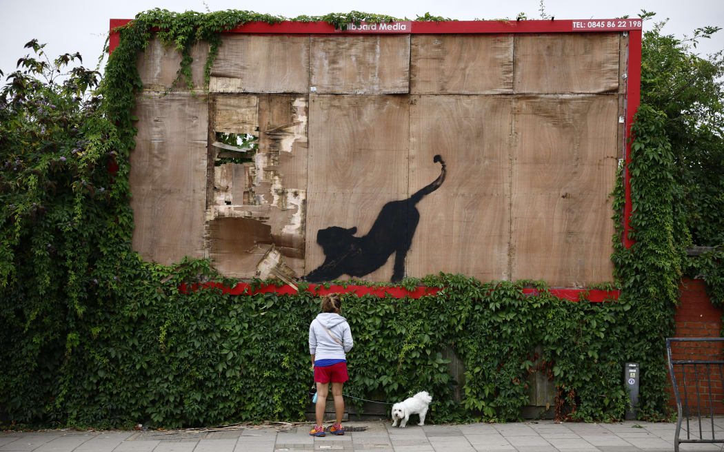 A pedestrian stops to look at an artwork by street artist Banksy, the sixth to be released this week, depicting a big cat looking up toward a hole in a disused advertising billboard, in northwest London, on August 10, 2024. The artist confirmed the work to be theirs after posting an image of it on the social media app Instagram. (Photo by BENJAMIN CREMEL / AFP) / RESTRICTED TO EDITORIAL USE - MANDATORY MENTION OF THE ARTIST UPON PUBLICATION - TO ILLUSTRATE THE EVENT AS SPECIFIED IN THE CAPTION