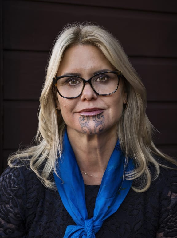 Hinemoa looks at the camera. She is wearing a black top with a blue scarf and standing against a dark background.