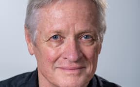 Opera director John Davies looks at the camera. He stands in front of a grey background. John looks like he's in his late 50s. His main feature is piercing blue eyes that almost feel as though they are looking through the camera and into your soul.
