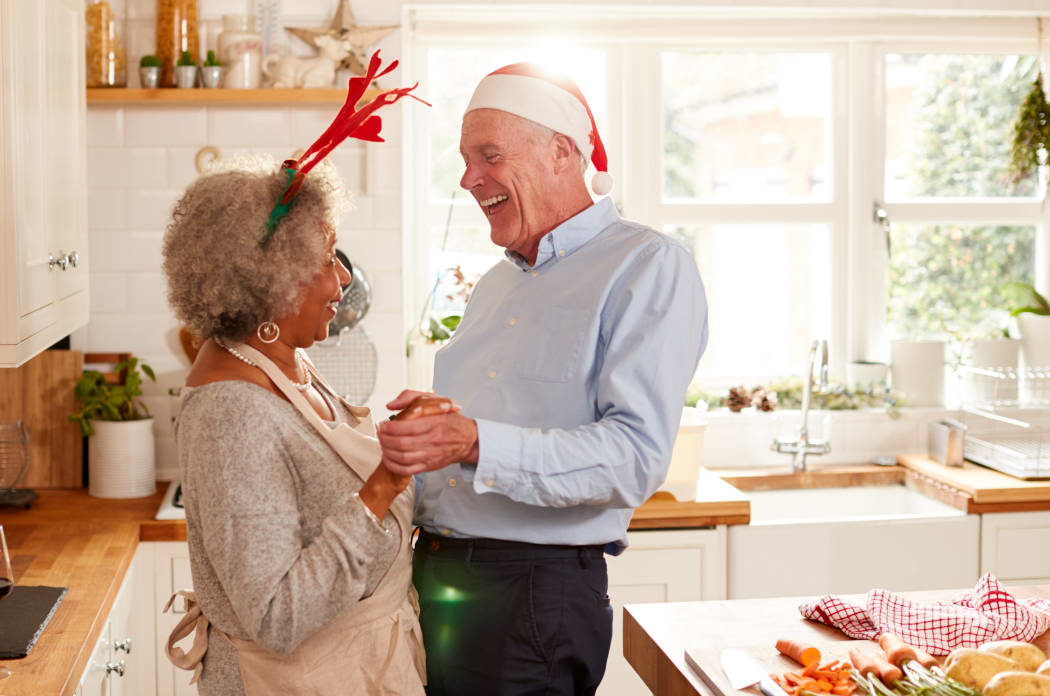 Loving Senior Couple Wearing Fancy Dress Antlers Dance In Kitchen Whilst Preparing Christmas Dinner