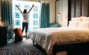 A woman backpacker stands next to her luggage and a window in a hotel room.