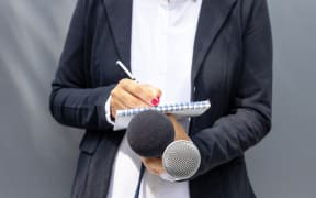 Female journalist at a news conference, writing notes, holding microphone.