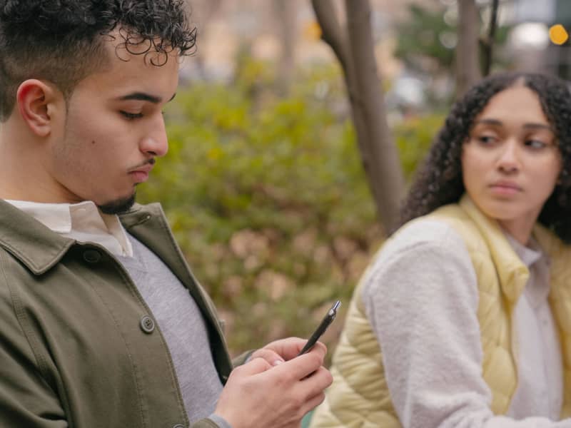 A young man looks at his phone while a young woman looks at him.