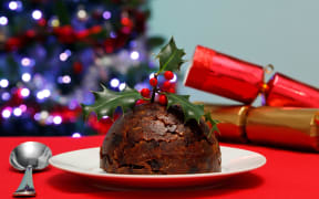 A photo of a Christmas pudding with holly on top with tree and crackers in the background.