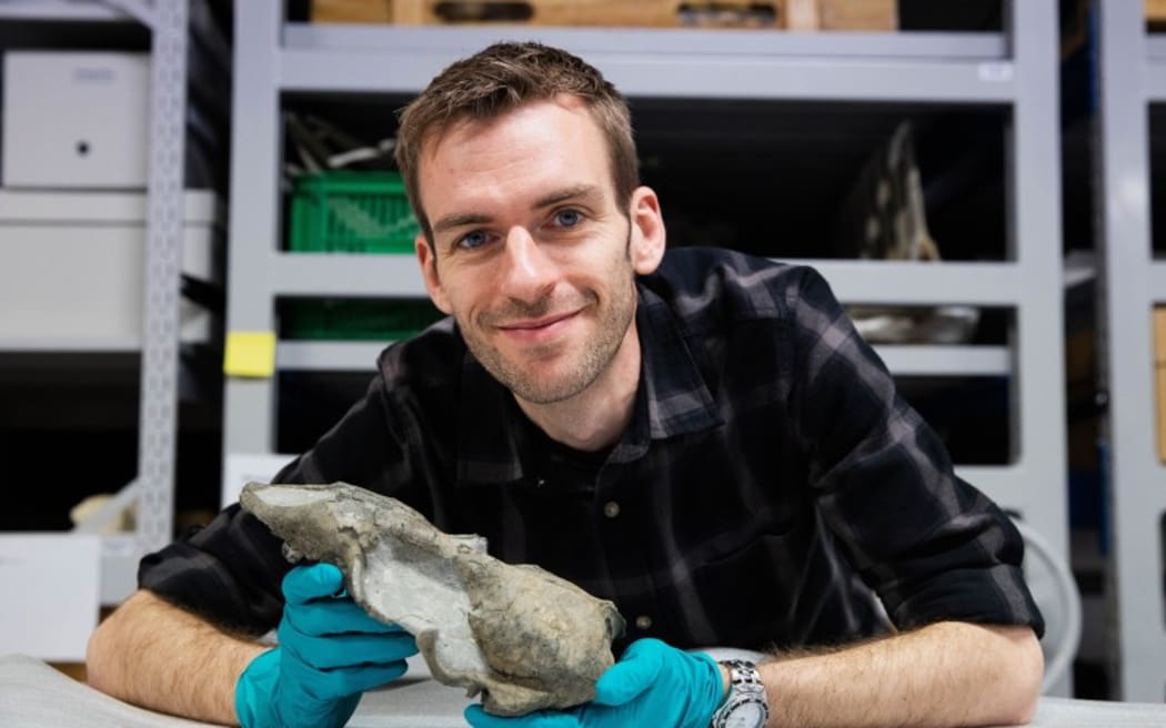 Felix Marx is wearing a pair of blue latex gloves, and holding what looks like a bone. He is leaning forwards over a table and smiling at the camera.
