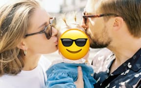 Parents holding a baby up, with an emoji covering the baby's face.