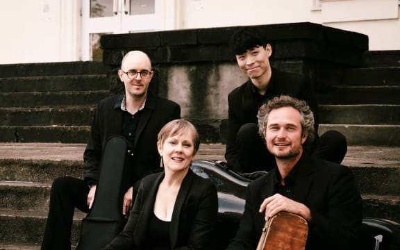 Akarana Piano Quartet members smiling, dressed in black, sit outside on some steps with their instruments in travel cases (but  no piano).