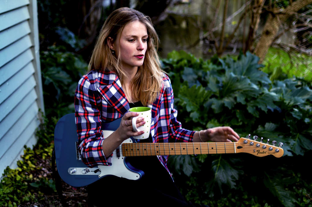 A colour photo of a young Women sitting down holding a guitar and a coffee cup