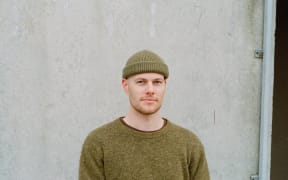 A man in a green beanie and top looks at the camera. He stands in front of a white wall.