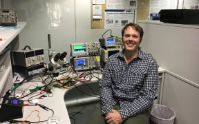 Wireless power engineer Daniel McCormick sits next to a lab bench with electronic equipment and a microscope.