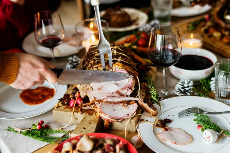 A person slicing roasted ham on a dinner table full of food plates and wine.