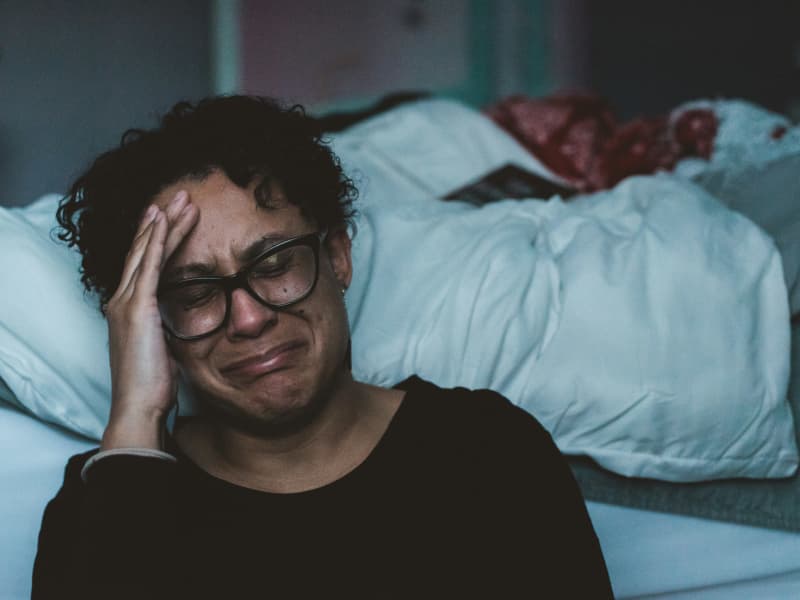 A woman with short hair and black-rimmed glasses cries on the edge of an unmade bed.
