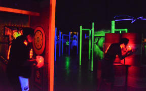 Two people are crouched in action in a laser tag arena. All the lights are out, and walls and weapons are lit up with fluorescent colours.