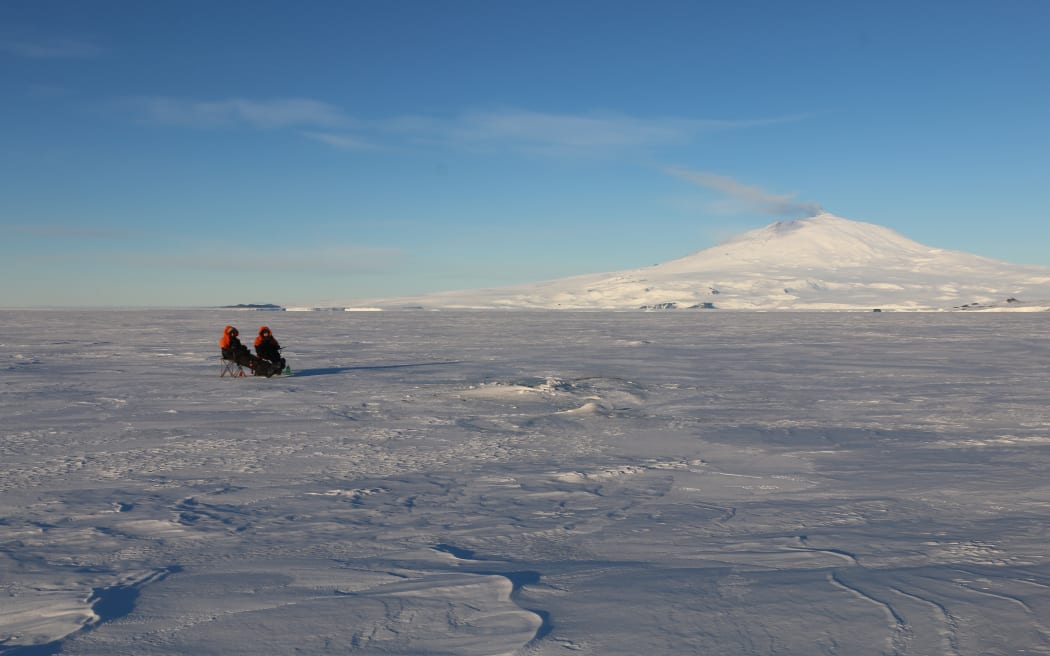 A wide shot: in the mid-distance, two people wearing black-and-orange sit on camp chairs on a wide, flat expanse of white ice with a snow-covered volcano on the horizon, and a blue sky above.