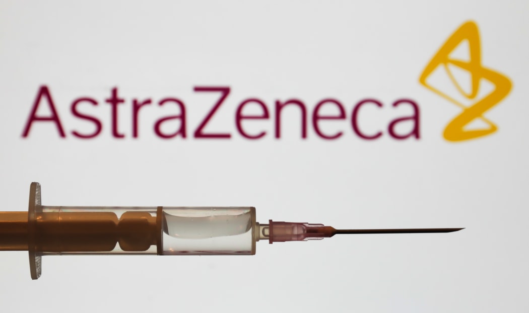 Medical syringe is seen with AstraZeneca company logo displayed on a screen in the background.
