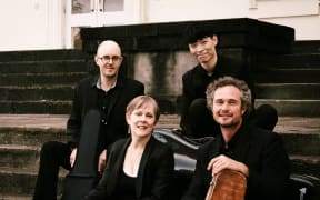 Akarana Piano Quartet members smiling, dressed in black, sit outside on some steps with their instruments in travel cases (but  no piano).
