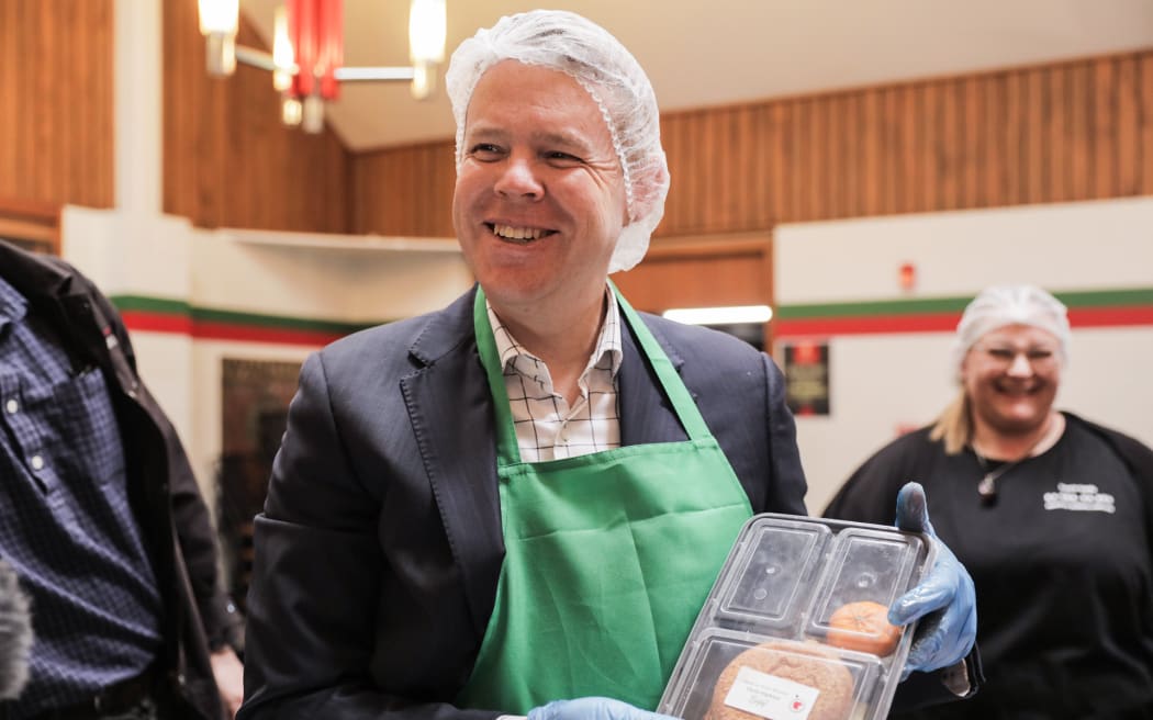 Chris Hipkins is visiting the Post Office Hotel in Pahiatua. They make free school lunches for approx 900 students in the area. Hipkins is helping make burgers which will be delivered to schools later.