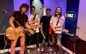 The four members of Voom with their respective instruments against a teal and white sound-paneled wall. They are lit with a purple light from behind them.